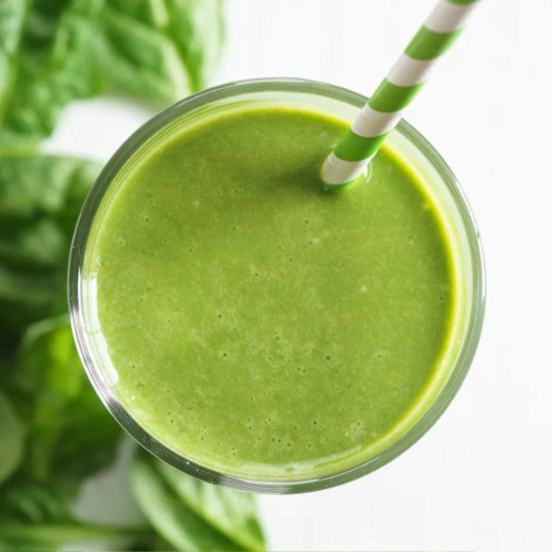 A glass filled with a smooth, vibrant green smoothie and a green and white striped straw, surrounded by fresh spinach leaves on a white background.